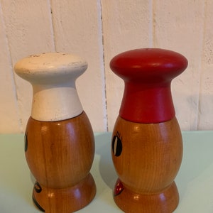 May include: A set of two wooden salt and pepper shakers with painted faces. The salt shaker has a white chef's hat and the pepper shaker has a red chef's hat.