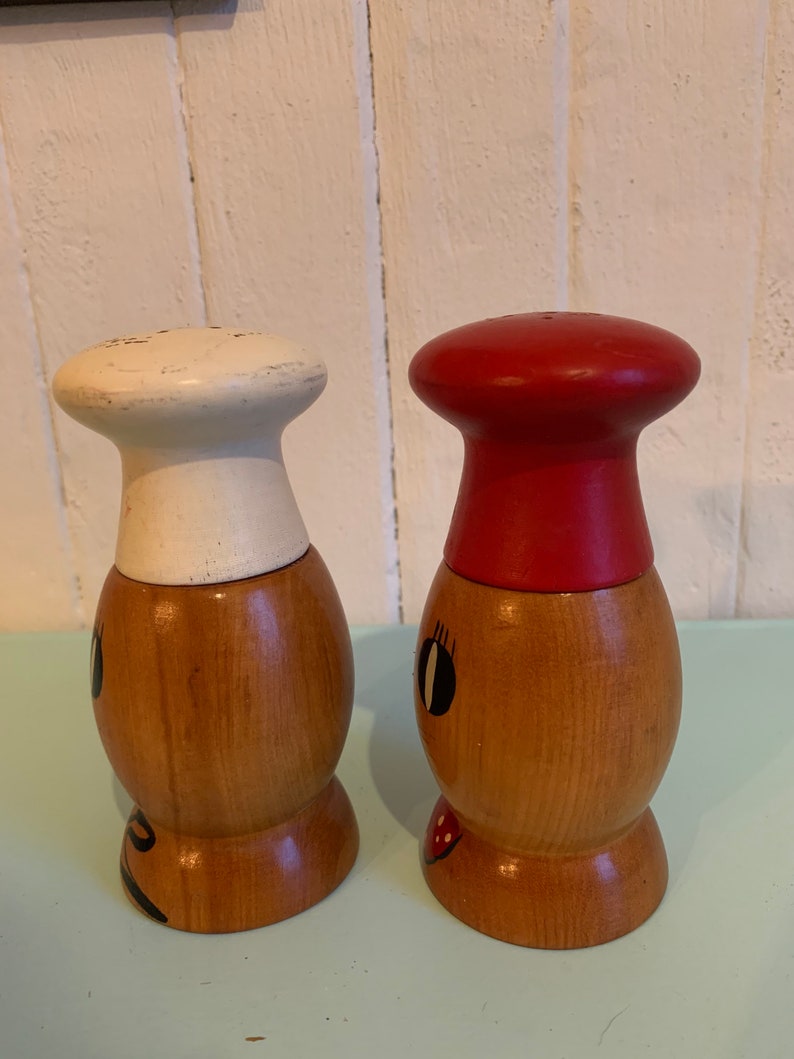 May include: A set of two wooden salt and pepper shakers with painted faces. The salt shaker has a white chef's hat and the pepper shaker has a red chef's hat.