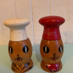 May include: A pair of wooden salt and pepper shakers shaped like cartoon chefs. The salt shaker has a white chef's hat and the pepper shaker has a red chef's hat. Both shakers have painted brown faces with black eyes and a red nose.