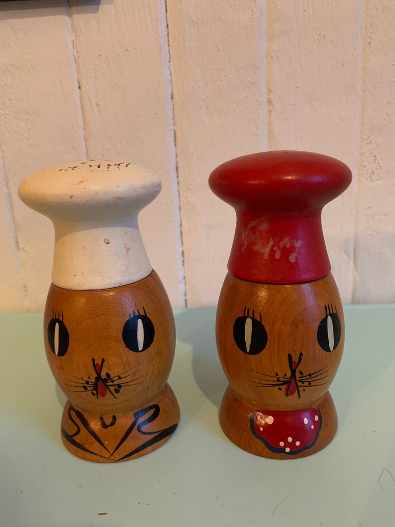 May include: A pair of wooden salt and pepper shakers shaped like cartoon chefs. The salt shaker has a white chef's hat and the pepper shaker has a red chef's hat. Both shakers have painted brown faces with black eyes and a red nose.