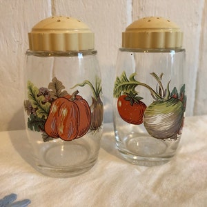 Peut inclure: Deux salières et poivrières en verre avec des couvercles crème et un motif de légumes. La salière de gauche a une citrouille, un oignon et des feuilles. La salière de droite a une tomate, un navet et des feuilles.