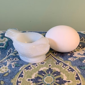 Peut inclure: Un œuf en céramique blanc en forme d'oiseau, avec un œuf blanc à côté. L'œuf et l'œuf sont sur un tissu à motifs bleus et verts.