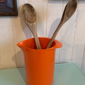 Könnte beinhalten: Zwei Holzlöffel befinden sich in einem orangefarbenen Plastikkrug. Der Krug hat eine breite Öffnung und einen Griff an einer Seite.