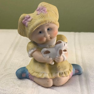 May include: A porcelain figurine of a little girl with blonde hair wearing a yellow dress and blue shoes. She is holding a white porcelain puppy in her arms.