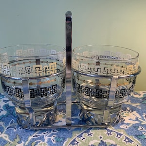 Peut inclure: Un ensemble de deux verres à boire en verre transparent avec un motif géométrique noir et or, dans un porte-verre en fil de chrome.