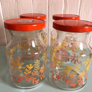 Puede incluir: Conjunto de cuatro tarros de almacenamiento de vidrio con tapas naranjas y dise&ntilde;os florales en tonos naranja y amarillo. Los tarros tienen un aspecto vintage y son perfectos para guardar especias, t&eacute; u otros art&iacute;culos de despensa.