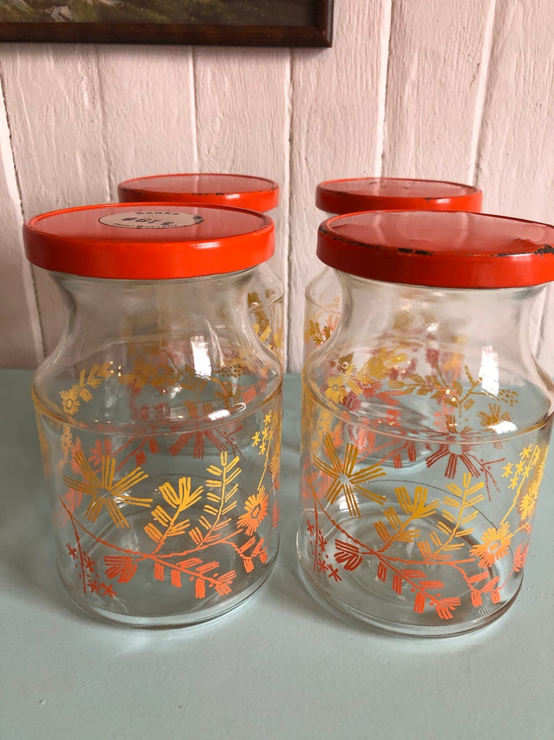 Puede incluir: Conjunto de cuatro tarros de almacenamiento de vidrio con tapas naranjas y dise&ntilde;os florales en tonos naranja y amarillo. Los tarros tienen un aspecto vintage y son perfectos para guardar especias, t&eacute; u otros art&iacute;culos de despensa.