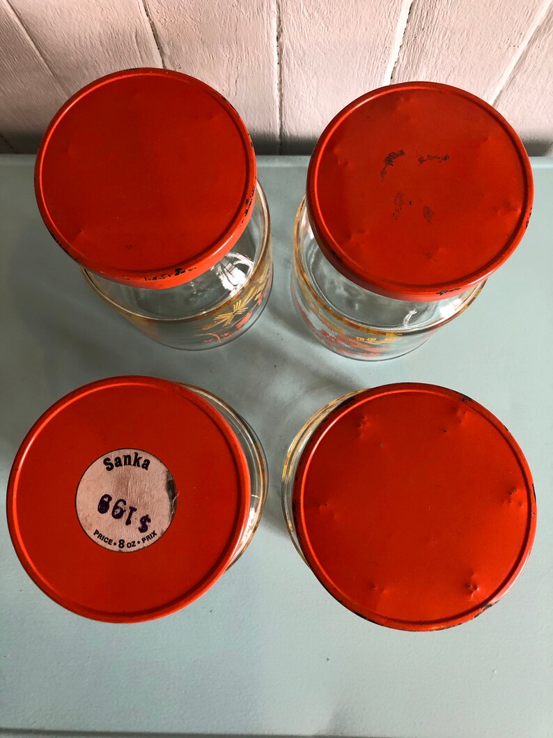 Puede incluir: Cuatro tarros de vidrio naranja con tapas. Los tarros tienen un dise&ntilde;o floral y las tapas son de un naranja brillante. Un tarro tiene una etiqueta que dice "Sanka 86&cent; PRICE: 8 OZ. - PRIX"