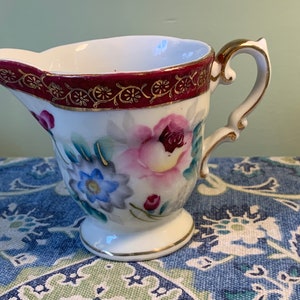 Puede incluir: Un pequeño creamer blanco con un asa dorada y un diseño floral. El creamer tiene una banda burdeos con detalles dorados alrededor del borde.