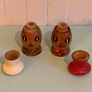 May include: A set of two wooden salt and pepper shakers with painted faces. The shakers are shaped like eggs and have a brown base with a white top. The faces are painted with black eyes, a black nose, and a red mouth. The shakers are sitting on a light blue surface.