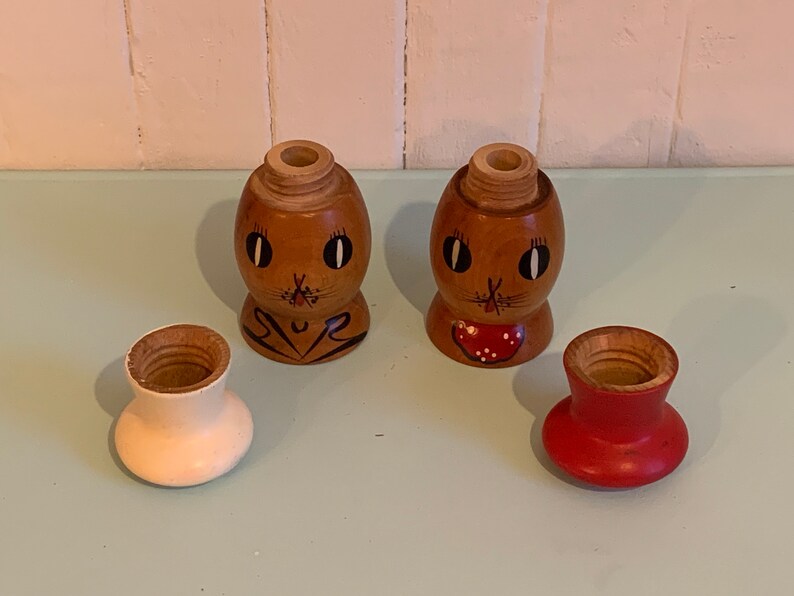 May include: A set of two wooden salt and pepper shakers with painted faces. The shakers are shaped like eggs and have a brown base with a white top. The faces are painted with black eyes, a black nose, and a red mouth. The shakers are sitting on a light blue surface.