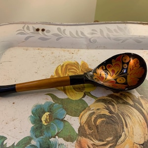 Puede incluir: Una cuchara de madera con mango negro y cuenco decorado con diseño floral en rojo, naranja y dorado. La cuchara está sobre una bandeja con estampado floral sobre fondo blanco.