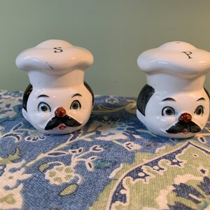 Peut inclure: Un ensemble de deux salières et poivrières en céramique blanche en forme de chefs avec des cheveux noirs, des yeux bruns, des nez rouges et des moustaches noires. Les salières ont des toques de chef blanches avec les lettres "S" et "P" dessus.