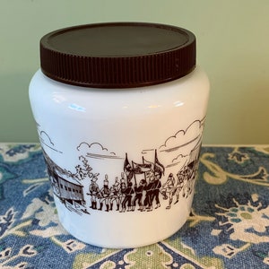 Peut inclure: Pot en céramique blanche avec un couvercle brun et une illustration brune d'un groupe de personnes en parade. L'illustration comprend un bâtiment, des arbres et des drapeaux.