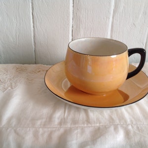 Puede incluir: Un juego de taza y platillo vintage con un esmalte naranja pálido y un borde negro. La taza tiene una forma redondeada y un asa negra. El platillo es del mismo color y tiene un borde negro.