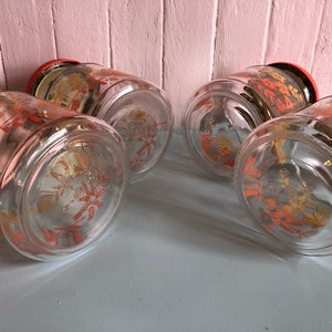 Puede incluir: Conjunto de cuatro tarros de almacenamiento de vidrio con tapas rojas y un dise&ntilde;o floral en naranja y amarillo. Los tarros est&aacute;n vac&iacute;os y tienen un cuerpo de vidrio transparente.