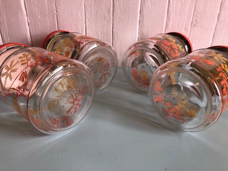 Puede incluir: Conjunto de cuatro tarros de almacenamiento de vidrio con tapas rojas y un dise&ntilde;o floral en naranja y amarillo. Los tarros est&aacute;n vac&iacute;os y tienen un cuerpo de vidrio transparente.