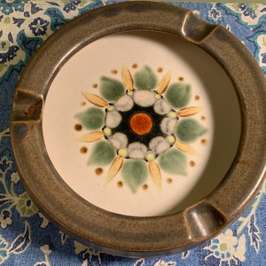 Peut inclure: Un cendrier en céramique brun avec un centre blanc et un motif floral coloré. Le motif présente des couleurs vertes, jaunes, oranges et noires.