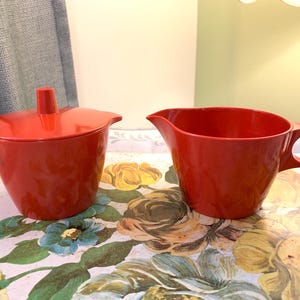 Peut inclure: Un sucrier rouge vintage avec un couvercle et une crémière rouge. Le sucrier a une poignée de chaque côté et un couvercle avec une petite poignée. La crémière a une poignée et un bec verseur. Les deux sont en matériau brillant.