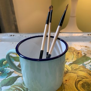 Op de afbeelding: Drie kwasten met houten handvatten en zwarte haren zitten in een lichtblauwe emaille mok met een donkerblauwe rand. De mok staat op een oppervlak met een bloemenpatroon.
