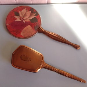 Peut inclure: Un ensemble de deux miroirs de poche vintage avec des poignées dorées. Un miroir est rond avec un motif floral rouge et blanc. L'autre miroir est rectangulaire avec une surface lisse et dorée.