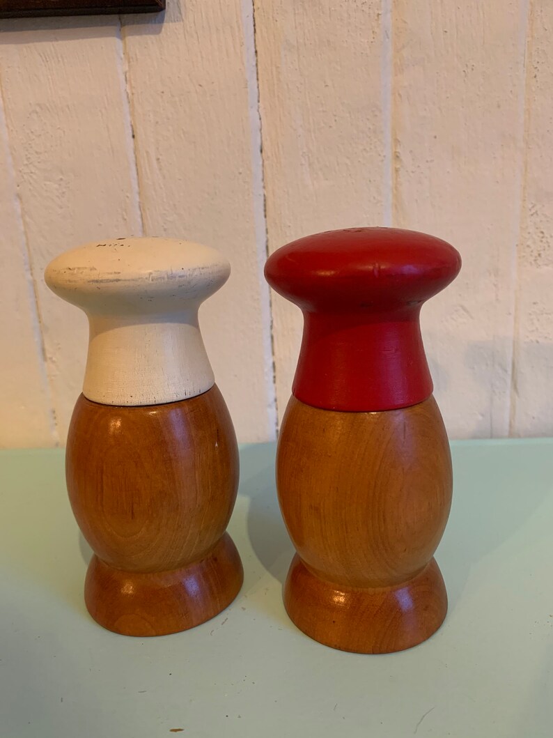 May include: A set of two wooden salt and pepper shakers. The shakers are shaped like a chef's hat. One shaker is painted white and the other is painted red.