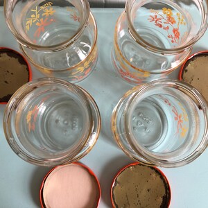 Puede incluir: Conjunto de cuatro tarros de vidrio con dise&ntilde;os florales naranja y amarillo y tapas naranjas. Los tarros est&aacute;n vac&iacute;os y las tapas est&aacute;n quitadas.