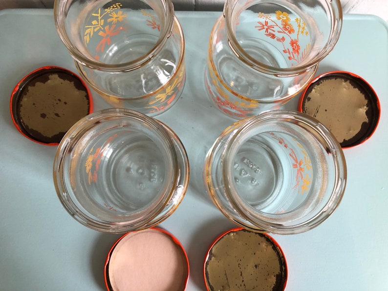 Puede incluir: Conjunto de cuatro tarros de vidrio con dise&ntilde;os florales naranja y amarillo y tapas naranjas. Los tarros est&aacute;n vac&iacute;os y las tapas est&aacute;n quitadas.