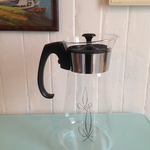 Può includere: Una caraffa di vetro trasparente con manico nero e coperchio argentato. La caraffa ha un design decorativo sul davanti.