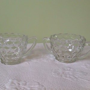 Könnte beinhalten: Ein Set aus zwei klaren Zucker- und Sahnebehältern aus Glas mit einem Diamantenmuster. Die Behälter haben einen einzelnen Griff.