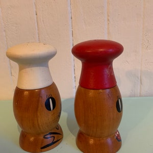 May include: A set of two wooden salt and pepper shakers shaped like chefs. The salt shaker has a white chef's hat and the pepper shaker has a red chef's hat. Both shakers have painted faces with black eyes and a red mouth.