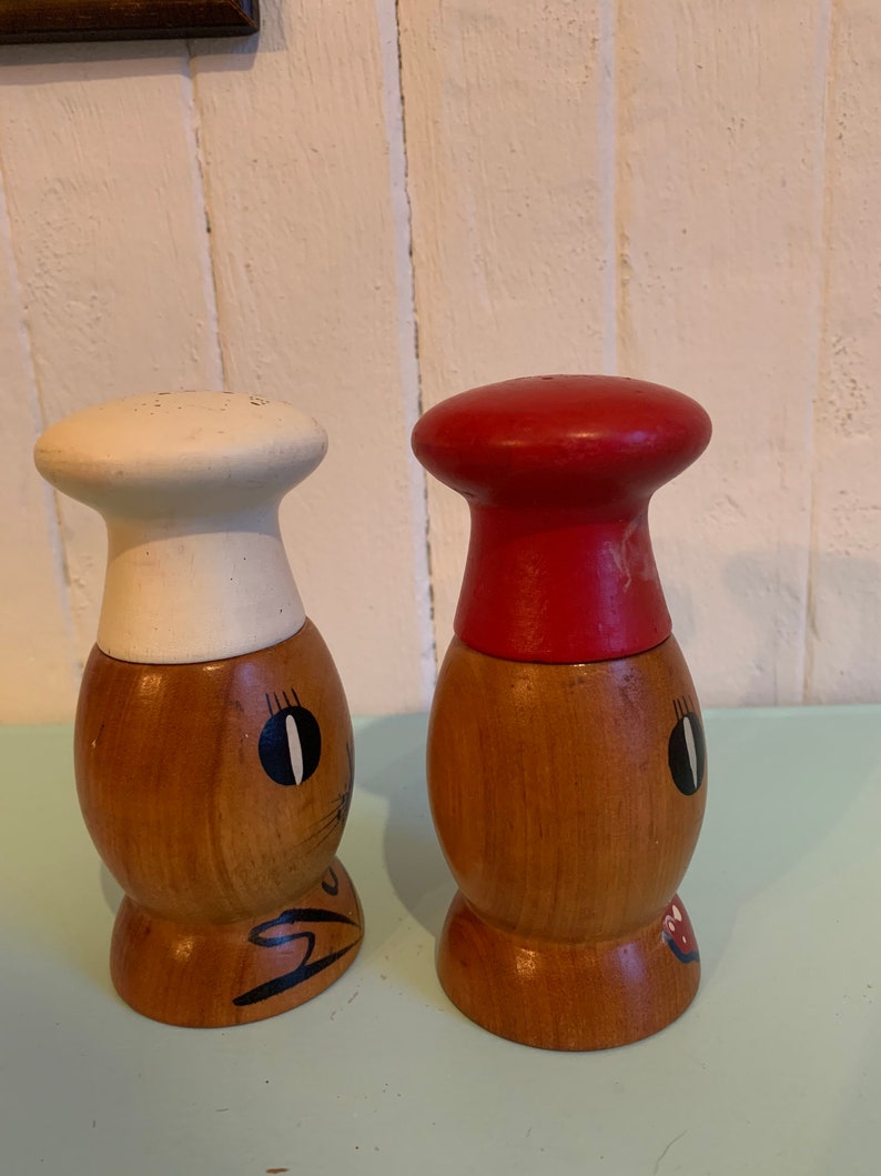 May include: A set of two wooden salt and pepper shakers shaped like chefs. The salt shaker has a white chef's hat and the pepper shaker has a red chef's hat. Both shakers have painted faces with black eyes and a red mouth.