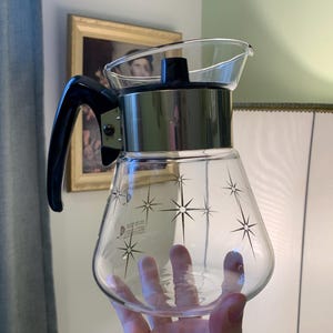 Op de afbeelding: Een glazen koffiekan met een zilveren deksel en een zwarte handgreep. De kan heeft een stervormig ontwerp aan de zijkant.