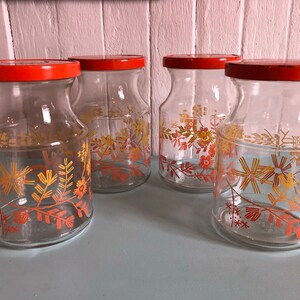 Puede incluir: Cuatro tarros de vidrio con tapas naranjas y dise&ntilde;os florales en tonos naranja y amarillo. Los tarros est&aacute;n sobre una superficie azul claro.