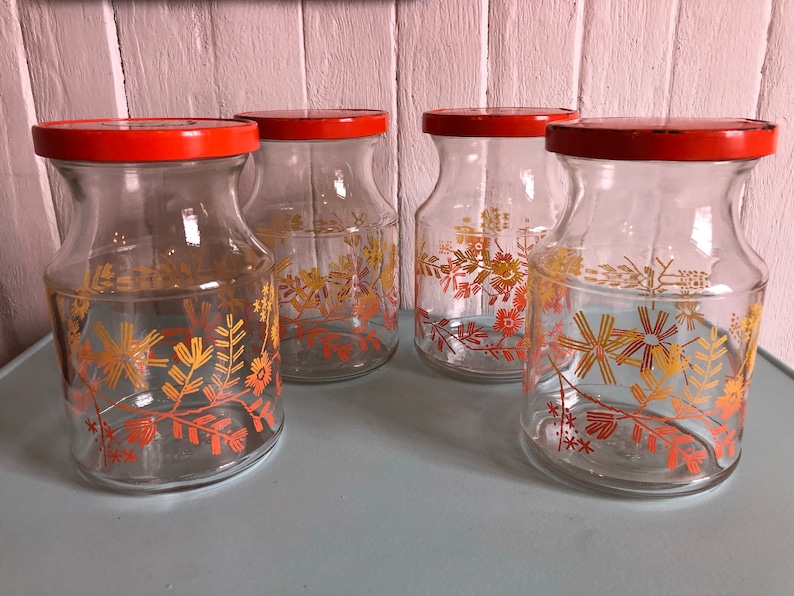 Puede incluir: Cuatro tarros de vidrio con tapas naranjas y dise&ntilde;os florales en tonos naranja y amarillo. Los tarros est&aacute;n sobre una superficie azul claro.