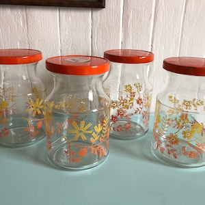 Peut inclure: Quatre bocaux en verre avec des couvercles orange et des motifs floraux dans des tons orange et jaune. Les bocaux sont vides et semblent être vintage.