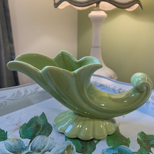 Könnte beinhalten: Eine hellgrüne Keramikvase in Hornform mit geriffeltem Sockel. Die Vase steht auf einer Oberfläche mit Blumenmuster.