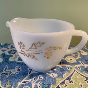 Puede incluir: Crema de vidrio de leche blanco con un diseño floral dorado. La crema tiene un asa y un pico de vertido.