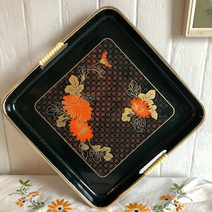 Könnte beinhalten: Ein quadratisches Serviertablett mit dunkelgrünem Hintergrund und goldenem Rand. Das Tablett hat ein rautenförmiges Mitteldesign mit orangefarbenen Blumenmustern und goldenen Akzenten. Es hat cremefarbene Griffe auf jeder Seite.