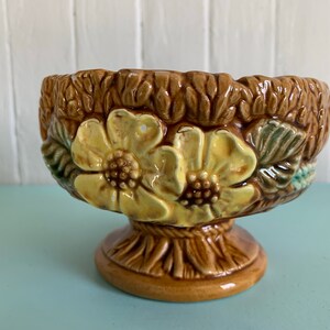 Peut inclure: Un pot en céramique brun avec une base en piédestal. Le pot est décoré de fleurs jaunes et de feuilles vertes.