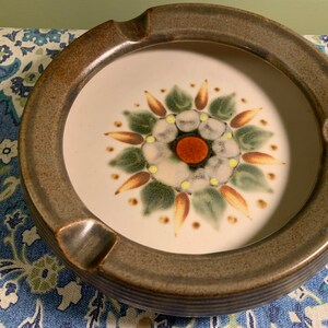 Peut inclure: Un cendrier en céramique brun avec un centre blanc orné d'un motif floral stylisé avec des accents verts, orange et jaunes.