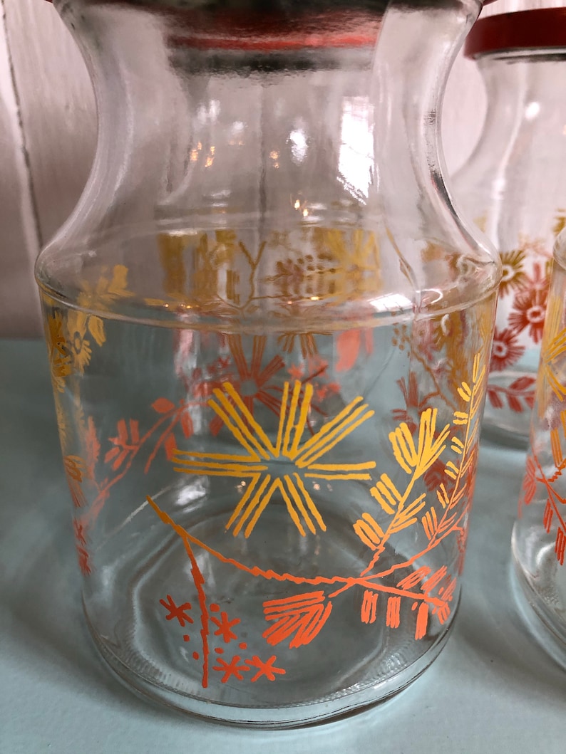 Puede incluir: Conjunto de cuatro tarros de vidrio transparente con tapas rojas y un dise&ntilde;o floral en naranja y amarillo. Los tarros son perfectos para guardar especias, hierbas u otros art&iacute;culos esenciales de cocina.