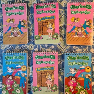 Peut inclure: Six mini-carnets Cabbage Patch Kids avec des couvertures roses, bleues et vertes. Chaque carnet présente une illustration différente des poupées Cabbage Patch Kids et le texte "Cabbage Patch Kids" et "Ptits Bouts de Choux".