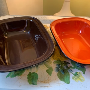 Peut inclure: Deux plats à four en émail, l'un brun et l'autre orange, avec des bords noirs. Les plats sont posés sur une surface blanche à motif floral.