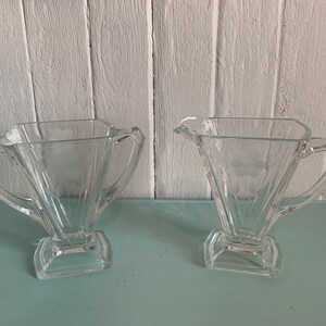 Peut inclure: Ensemble sucrier et crémier en verre transparent avec un motif géométrique. Les pièces ont une base carrée et un sommet triangulaire évasé.