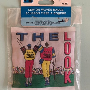 Peut inclure: Écusson tissé à coudre avec le texte "THE LOOK" en lettres colorées. Deux joueurs de baseball sont représentés au centre, portant des uniformes avec les numéros 36 et 38. L'écusson est emballé dans un sac en plastique transparent.