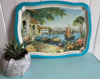 Può includere: Un vassoio vintage in metallo blu e bianco con una scena dipinta di un villaggio mediterraneo con barche sull'acqua. Il vassoio ha un bordo a festoni.