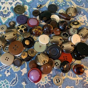 May include: A collection of assorted buttons in various colours, materials, and sizes. The buttons are made of wood, plastic, metal, and fabric. Some buttons have a four-hole design, while others have two holes. The buttons are arranged in a random pattern on a blue and white floral fabric.