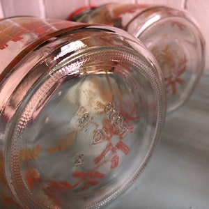 Puede incluir: Primer plano de la parte inferior de un tarro de vidrio transparente con un dise&ntilde;o floral rojo y naranja. El tarro tiene una superficie texturizada y un logotipo en relieve visible.