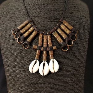 May include: A brown leather necklace with gold-toned metal accents and white shell pendants. The necklace is displayed on a brown mannequin.